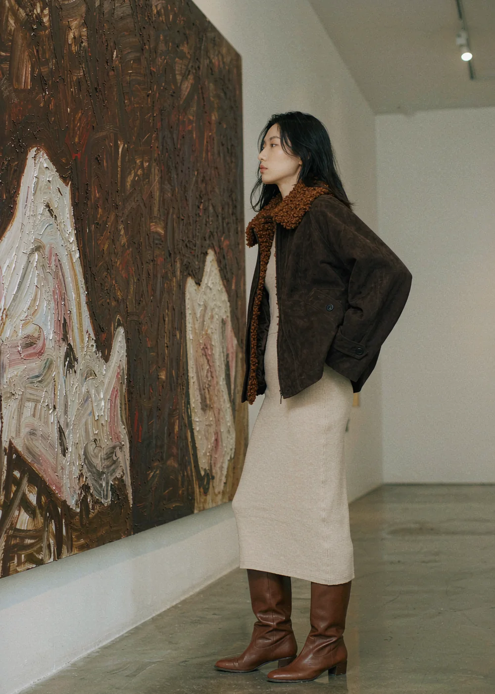 深棕麂皮夹克配奶油针织长裙与棕色长靴，站在画廊里